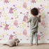 Lovely child standing in front of Whimsy Tots Cuddle Saurus Garden Wallpaper in Lilac Whisper Lavender with dinosaur, flower, bone, footprint in design.