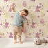 Lovely child standing in front of Whimsy Tots Cuddle Saurus Garden Wallpaper in Honey Beige with dinosaur, flower, bone, footprint in design.