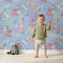 Lovely child standing in front of Whimsy Tots Cuddle Saurus Garden Wallpaper in Skyberry Blue with dinosaur, flower, bone, footprint in design.