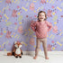 Lovely child standing in front of Whimsy Tots Cuddle Saurus Garden Wallpaper in Lavender Mist with dinosaur, flower, bone, footprint in design.