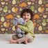 Lovely child standing in front of Whimsy Tots Hatchling Dino Garden Wallpaper in Berry Soil with dinosaur, egg, flower, leaf in design.