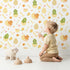 Lovely child standing in front of Whimsy Tots Hatchling Dino Parade Wallpaper in Soft Eggshell with dinosaur, egg, bone, flower in design.