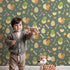 Lovely child standing in front of Whimsy Tots Hatchling Dino Parade Wallpaper in Olive Grove Green with dinosaur, egg, bone, flower in design.