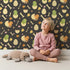 Lovely child standing in front of Whimsy Tots Hatchling Dino Parade Wallpaper in Charcoal Twilight Gray with dinosaur, egg, bone, flower in design.