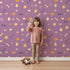 Lovely child standing in front of Whimsy Tots Dino Hatch Trail Wallpaper in Berry Bloom with egg, bone, flower, footprint in design.