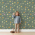 Lovely child standing in front of Whimsy Tots Dino Hatch Trail Wallpaper in Forest Fern Green with egg, bone, flower, footprint in design.