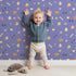Lovely child standing in front of Whimsy Tots Dino Hatch Trail Wallpaper in Lilac Sky Lavender with egg, bone, flower, footprint in design.