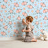Lovely child standing in front of Whimsy Tots Whispering Peony Garden Wallpaper in Sky Blossom with flower, peony, daisy, leaf in design.