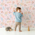 Lovely child standing in front of Whimsy Tots Floating Botanical Whispers Wallpaper in Blush Bloom Pink with flower, leaf, feather, peony in design.