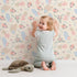 Lovely child standing in front of Whimsy Tots Floating Botanical Whispers Wallpaper in Cream Petal with flower, peony, leaf, feather in design.