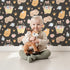 Lovely child standing in front of Whimsy Tots Blossom Basket Friends Wallpaper in Midnight Bloom with fox, mouse, flower, basket, crown in design.