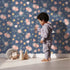 Lovely child standing in front of Whimsy Tots Twilight Garden Whimsy Wallpaper in Starry Blue with flower, leaf, star, bloom in design.