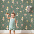 Lovely child standing in front of Whimsy Tots Blush Ballerina Dreams Wallpaper in Forest Waltz Green with ballerina, crown, tutu, slipper, flower in design.