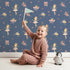 Lovely child standing in front of Whimsy Tots Blush Ballerina Dreams Wallpaper in Starlight Stage with ballerina, crown, tutu, slipper, flower in design.