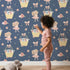 Lovely child standing in front of Whimsy Tots Ballet Mouse Garden Wallpaper in Twilight Bloom with mouse, flower, bow, basket in design.