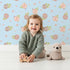 Lovely child standing in front of Whimsy Tots Soft Garden Posy Wallpaper in Sky Petal with flower, bouquet, leaf, heart in design.