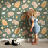 Lovely child standing in front of Whimsy Tots Royal Woodland Friends Wallpaper in Forest Crown Green with fox, rabbit, bear, flower, crown, star in design.