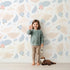 Lovely child standing in front of Whimsy Tots Whispering Petal Leaves Wallpaper in Cloud Cream with leaf, flower, petal in design.
