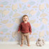 Lovely child standing in front of Whimsy Tots Whispering Petal Leaves Wallpaper in Lavender Mist with leaf, flower, petal in design.