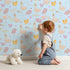 Lovely child standing in front of Whimsy Tots Crowned Swan Bloom Wallpaper in Sky Blue with swan, bow, crown, flower, leaf in design.