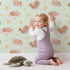 Lovely child standing in front of Whimsy Tots Woodland Nap Parade Wallpaper in Buttercream with bear, bunny, fox, flower, star in design.