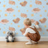 Lovely child standing in front of Whimsy Tots Woodland Nap Parade Wallpaper in Dreamy Blue with bear, bunny, fox, flower, star in design.