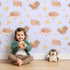 Lovely child standing in front of Whimsy Tots Woodland Nap Parade Wallpaper in Lavender Mist with bear, bunny, fox, flower, star in design.