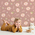 Lovely child standing in front of Whimsy Tots Blush Meadow Bloom Wallpaper in Clay Rose Brown with flower, daisy, leaf, petal in design.