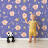 Lovely child standing in front of Whimsy Tots Blush Meadow Bloom Wallpaper in Lavender Bloom with flower, daisy, leaf, petal in design.