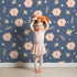 Lovely child standing in front of Whimsy Tots Blush Meadow Bloom Wallpaper in Midnight Blue with flower, daisy, leaf, petal in design.
