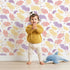 Lovely child standing in front of Whimsy Tots Pastel Feather Garden Wallpaper in Spring Mix with flower, leaf, feather, petal in design.