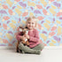 Lovely child standing in front of Whimsy Tots Pastel Feather Garden Wallpaper in Blue Skies with flower, leaf, feather, petal in design.