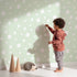Lovely child standing in front of Whimsy Tots Gentle Pebble Dots Wallpaper in Mint Cream with dot, spot, pebble, mint in design.
