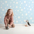Lovely child standing in front of Whimsy Tots Gentle Pebble Dots Wallpaper in Sky Blue with dot, spot, pebble, blue in design.