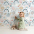 Lovely child standing in front of Whimsy Tots Pastel Bloom Rainbow Wallpaper in Sky Blue with rainbow, flower, leaf, petal, stripe in design.