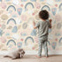 Lovely child standing in front of Whimsy Tots Pastel Bloom Rainbow Wallpaper in Cream with rainbow, flower, leaf, stem, arc in design.