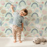Lovely child standing in front of Whimsy Tots Pastel Bloom Rainbow Wallpaper in Mint Green with rainbow, flower, arc, leaf, petal in design.