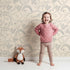 Lovely child standing in front of Whimsy Tots Doodle Meadow Shapes Wallpaper in Cream with doodle, shape, petal, scribble, arch in design.