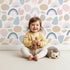 Lovely child standing in front of Whimsy Tots Rainbow Petal Shapes Wallpaper in White with rainbow, petal, arc, shape, stripe in design.