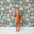 Lovely child standing in front of Whimsy Tots Rainbow Petal Shapes Wallpaper in Forest Green with rainbow, petal, shape, arch, stripe in design.