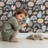Lovely child standing in front of Whimsy Tots Rainbow Petal Shapes Wallpaper in Charcoal Gray with rainbow, petal, shape, arch, stripe in design.