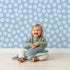 Lovely child standing in front of Whimsy Tots Playful Pebble Dots Wallpaper in Periwinkle Blue with dot, pebble, spot, oval in design.