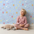 Lovely child standing in front of Whimsy Tots Cupids Arrow Parade Wallpaper in Sky Blue with arrow, heart, star in design.