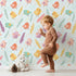 Lovely child standing in front of Whimsy Tots Feathered Joy Parade Wallpaper in Sky Mint with bird, feather, sprig, arrow in design.