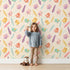 Lovely child standing in front of Whimsy Tots Feathered Joy Parade Wallpaper in Warm Cream with bird, feather, arrow, sprig in design.