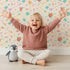 Lovely child standing in front of Whimsy Tots Love and Arrows Bloom Parade Wallpaper in Soft Peach with arrow, heart, flower, burst in design.