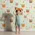 Lovely child standing in front of Whimsy Tots Woodland Tribe Hearts Wallpaper in Soft Mint Green with bear, fox, rabbit, feather, arrow, heart in design.