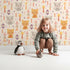 Lovely child standing in front of Whimsy Tots Mindful Kitten Heart Wallpaper in Oat Beige with cat, heart, triangle, dot, diamond in design.