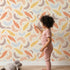 Lovely child standing in front of Whimsy Tots Feathered Whimsy Drift Wallpaper in Oat Beige with feather, diamond, dot in design.