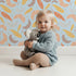 Lovely child standing in front of Whimsy Tots Feathered Whimsy Drift Wallpaper in Sky Blue with feather, diamond, dot in design.